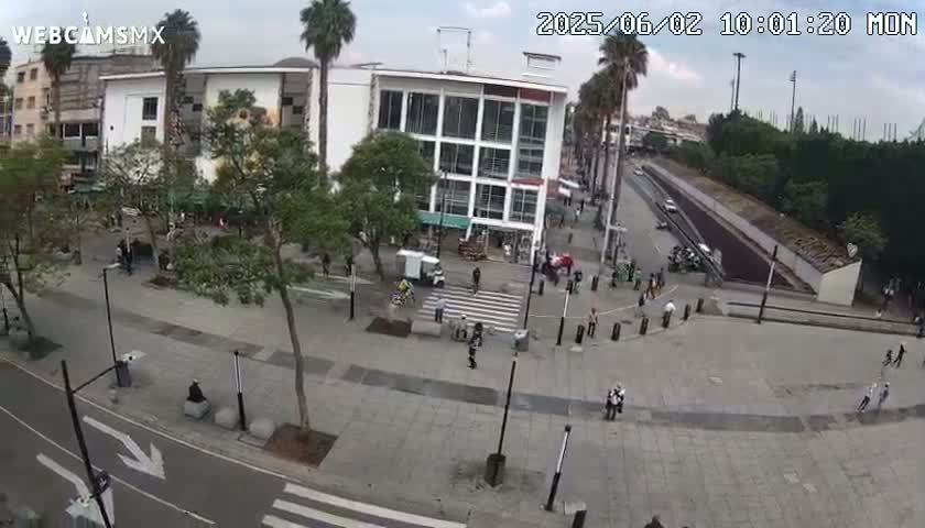 An aerial view of a city square on an overcast day, showing pedestrians walking around a multi-story building and a street with sparse traffic.