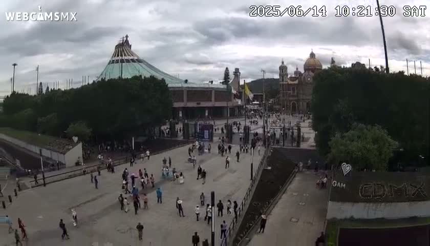 A large plaza with many people, surrounded by buildings including a large basilica and a modern structure under an overcast sky.