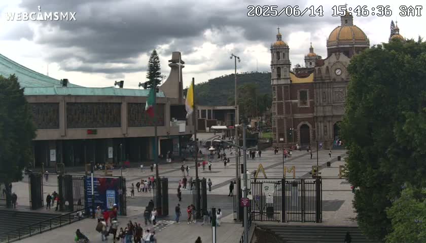 A large plaza with many people, surrounded by buildings including a large basilica and a modern structure under an overcast sky.