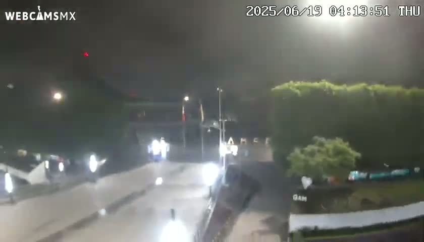 A nighttime, possibly rainy, scene shows a mostly empty road with some vehicles and buildings in the background.