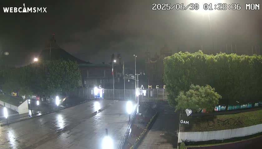 A nighttime view of a wet plaza and building complex under a cloudy sky, with streetlights illuminating the scene.