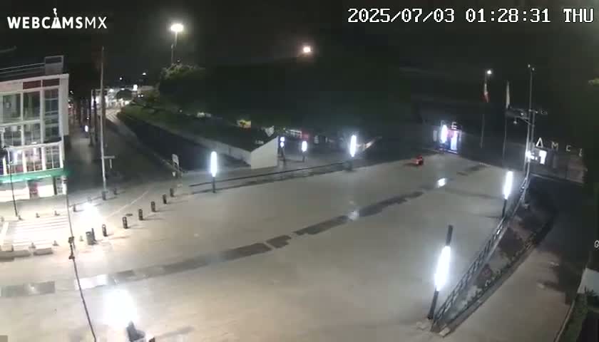 A mostly empty plaza at night, illuminated by several lights, is wet from a recent rain.