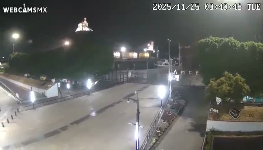 The image shows a wet, deserted urban area at night, illuminated by streetlights, with a prominent statue atop a distant building or hill under a dark, possibly overcast sky after rain.