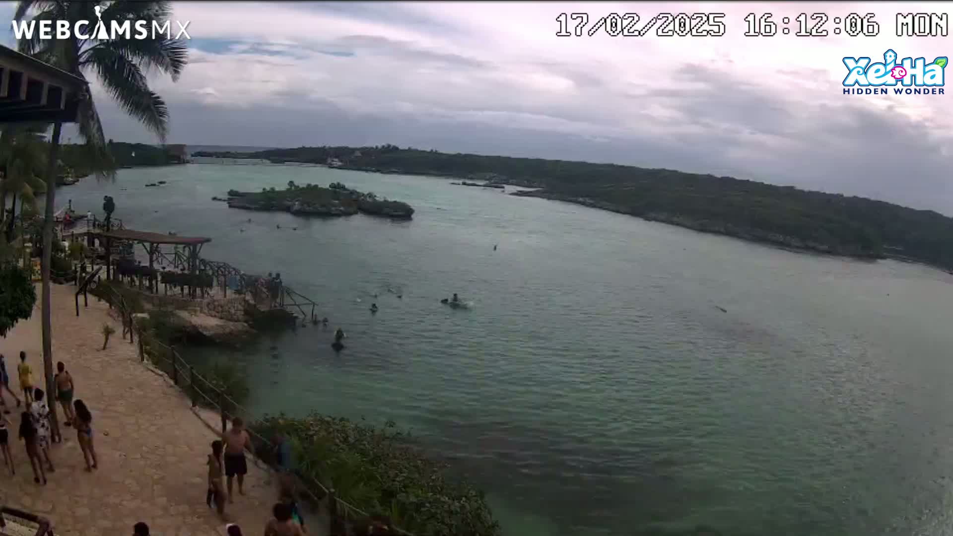 An overcast day shows a calm body of water with people swimming and relaxing near a rocky shore, where others are walking along a paved path.