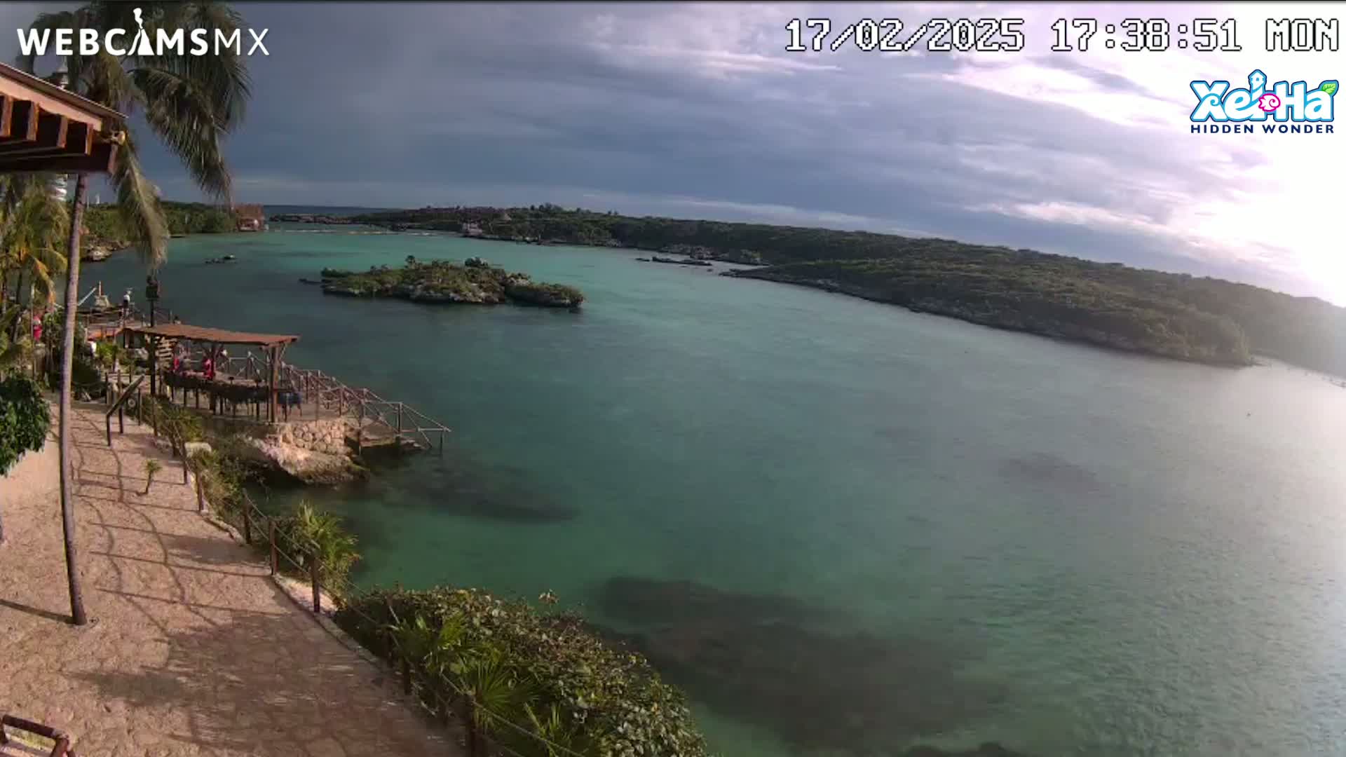 A partly cloudy day shows a tranquil bay of teal water, surrounded by lush green vegetation and a small rocky island, viewed from a high vantage point overlooking a sandy path and a wooden structure.