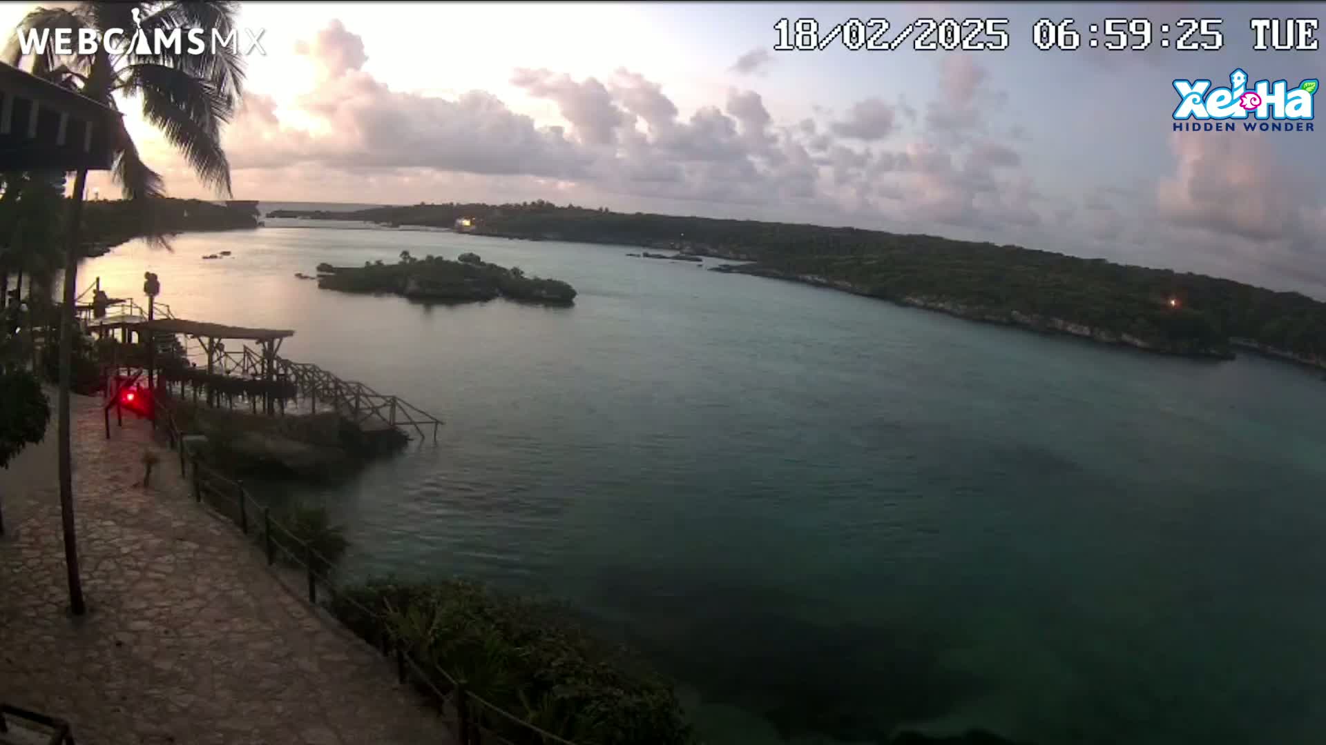 A tranquil sunrise scene shows a calm body of water surrounded by lush green land, with a wooden dock and stone pathway visible in the foreground.