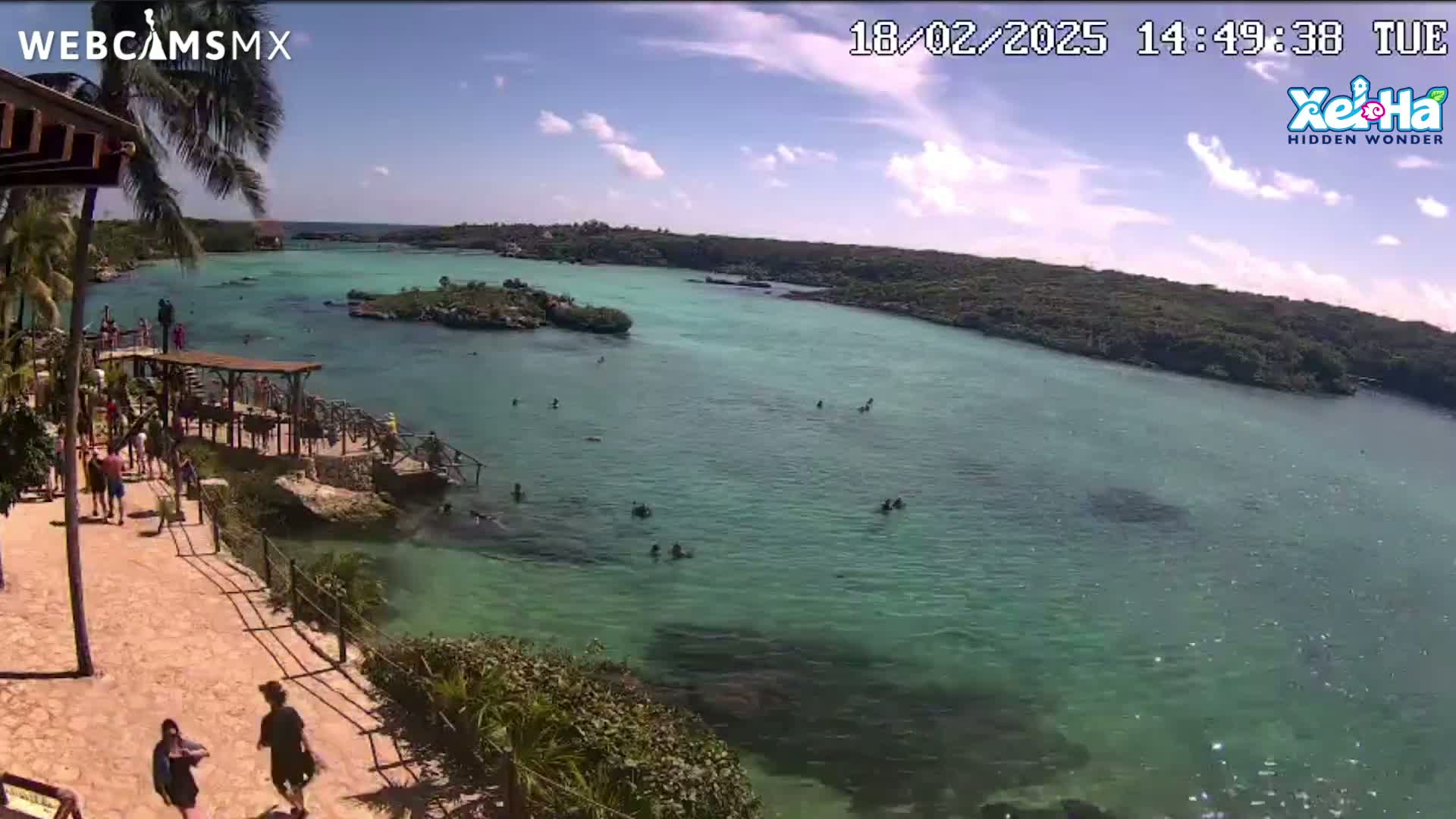 A partly sunny day shows a tropical lagoon with clear, teal water, people swimming and sunbathing, and a walkway leading to a dock from a stone pathway lined with vegetation.