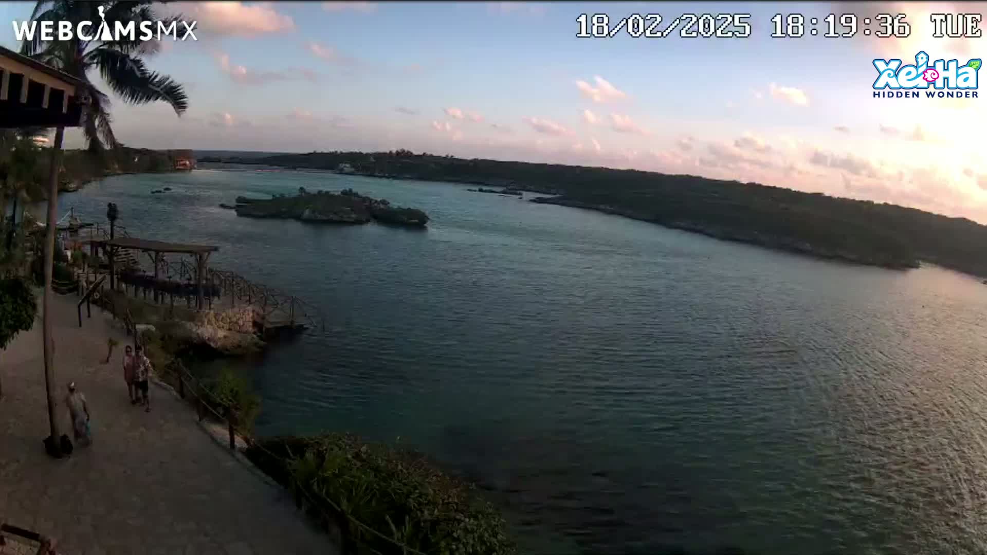 An elevated view shows a calm body of water at sunset, with a few people walking along a paved path near a rocky shoreline and some small islands in the distance.