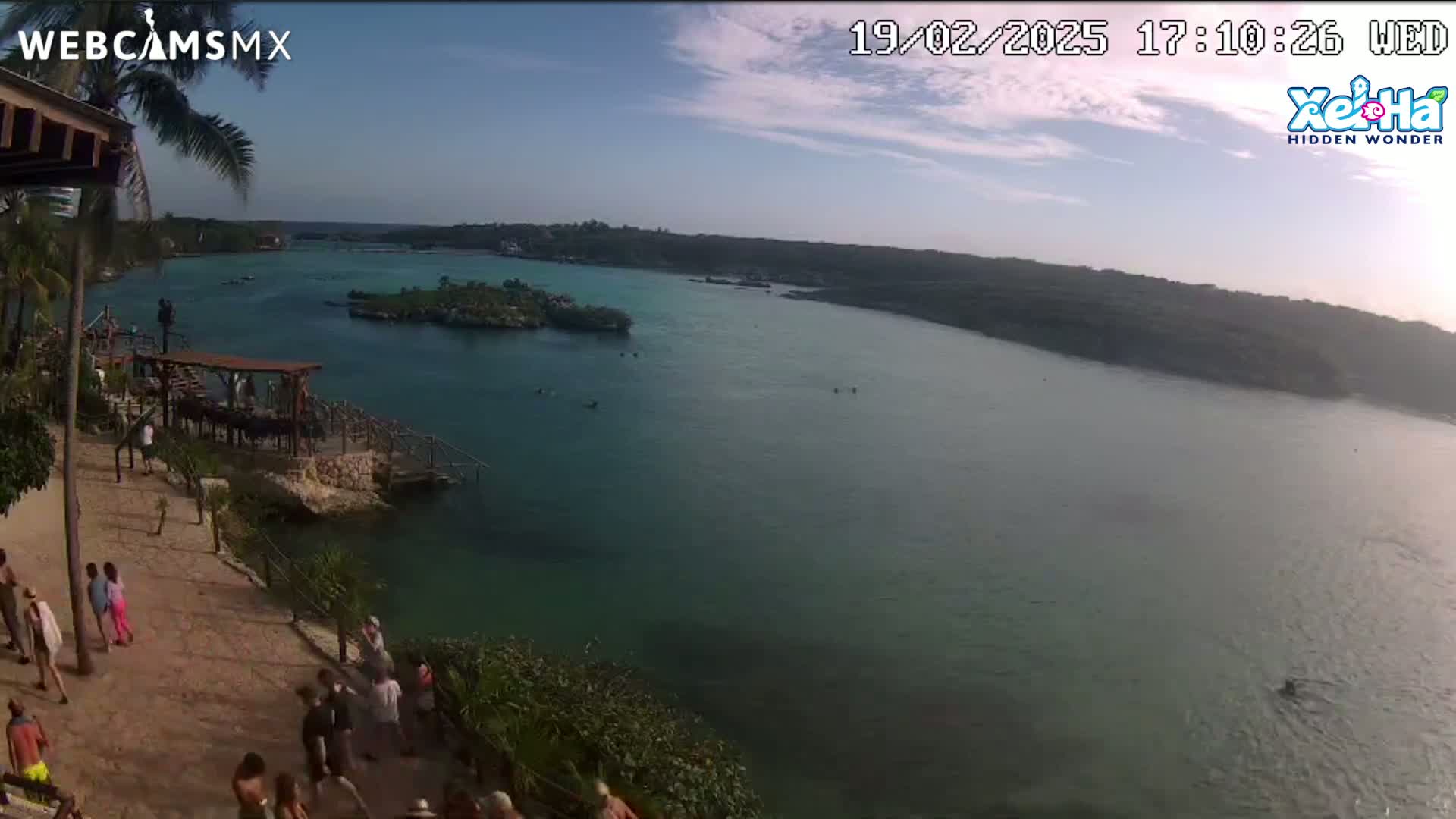 An elevated view overlooking a calm, turquoise body of water with small islands and a shoreline populated with people under a partly cloudy sky.