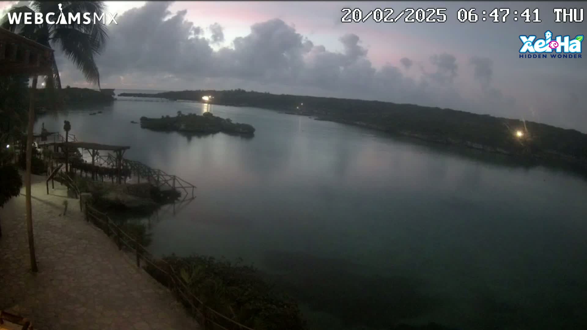 A calm body of water, partially shrouded in mist under a cloudy, dusky sky, is viewed from a high vantage point overlooking a stone pathway and wooden structures.