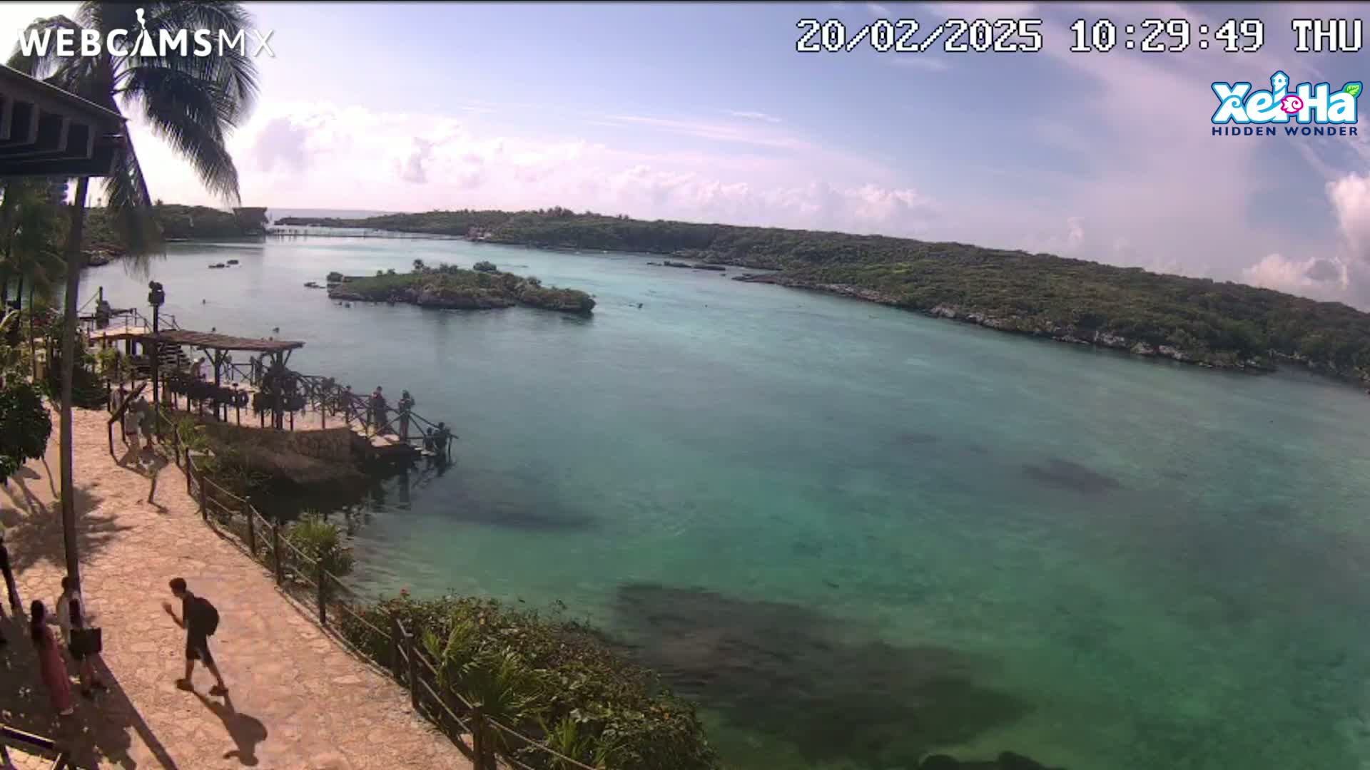 A partly sunny day shows a tranquil, teal-colored body of water surrounded by lush green vegetation, with people walking along a stone path near a dock.