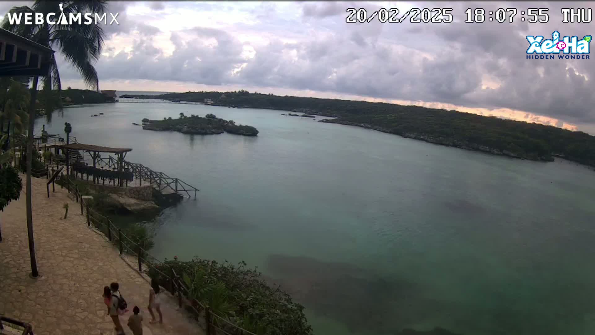 A tranquil cove with calm, greenish-blue water is viewed from a high vantage point, under a partly cloudy, sunset sky; several people walk along a stone path overlooking the water.