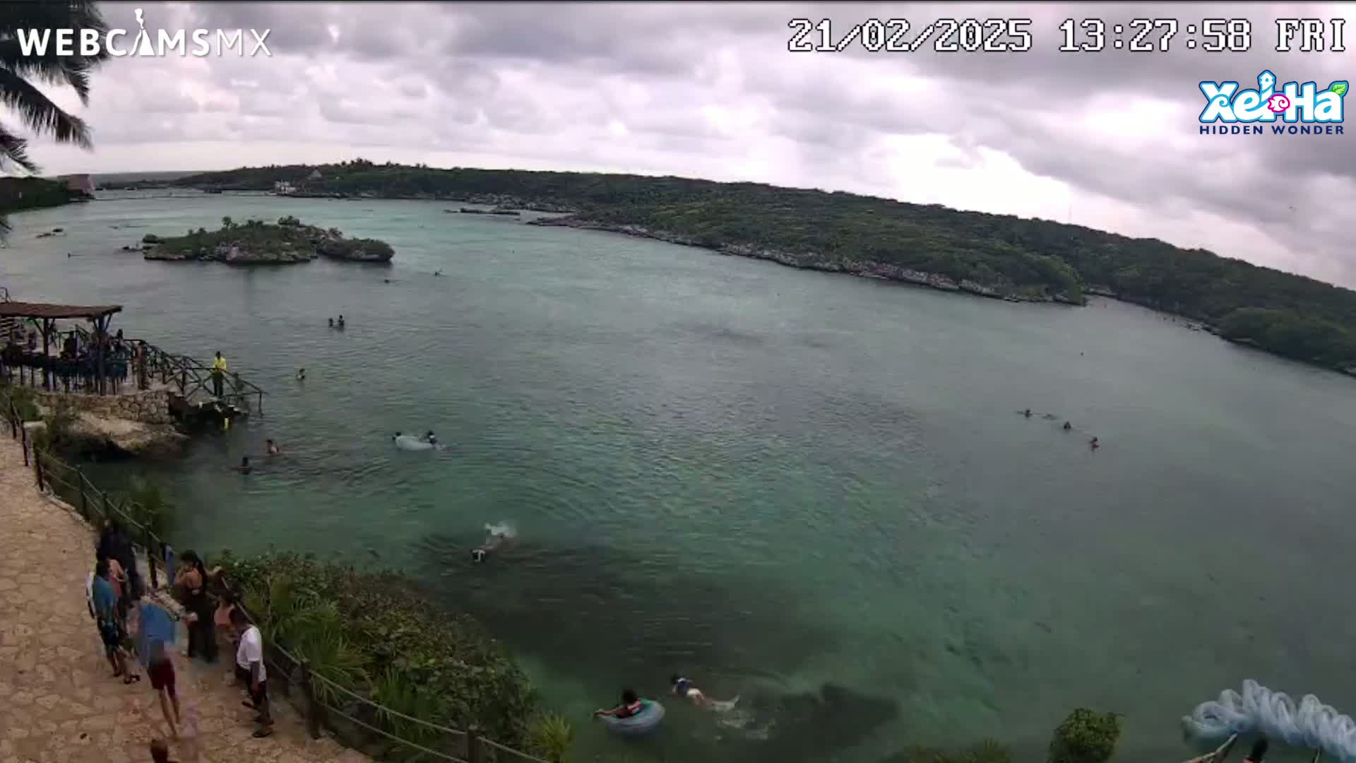 An overcast day at a calm, turquoise body of water shows people swimming and relaxing near a rocky shoreline with lush green vegetation.