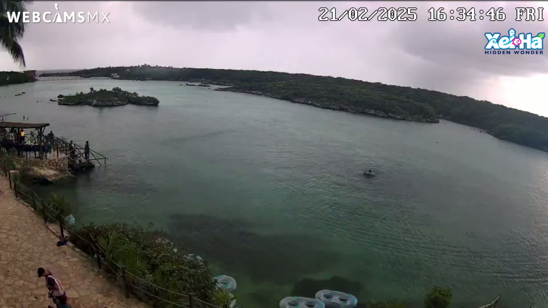 An overcast day shows a calm body of green water, surrounded by lush green vegetation, with a small island in the middle and several people near a dock.