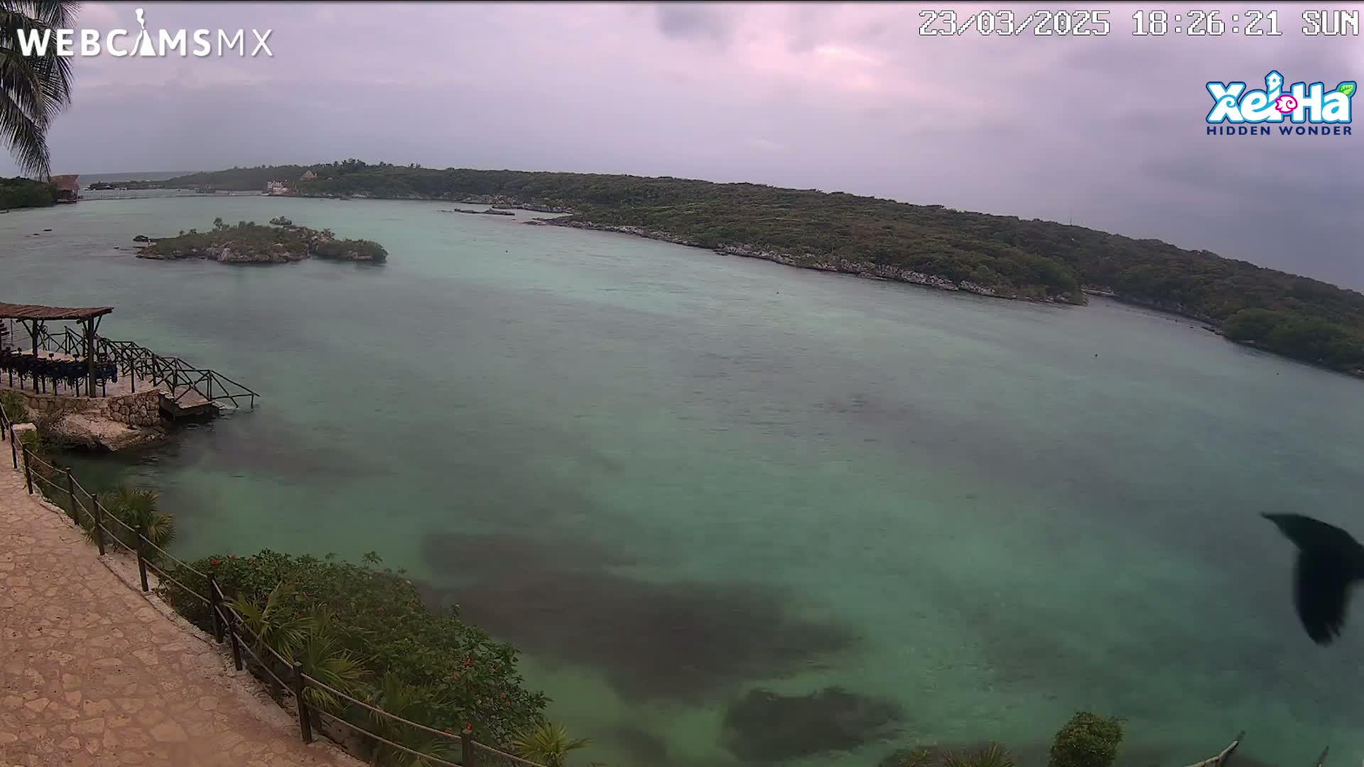 A rocky shoreline with a wooden structure overlooks a calm, teal-green body of water under an overcast sky.