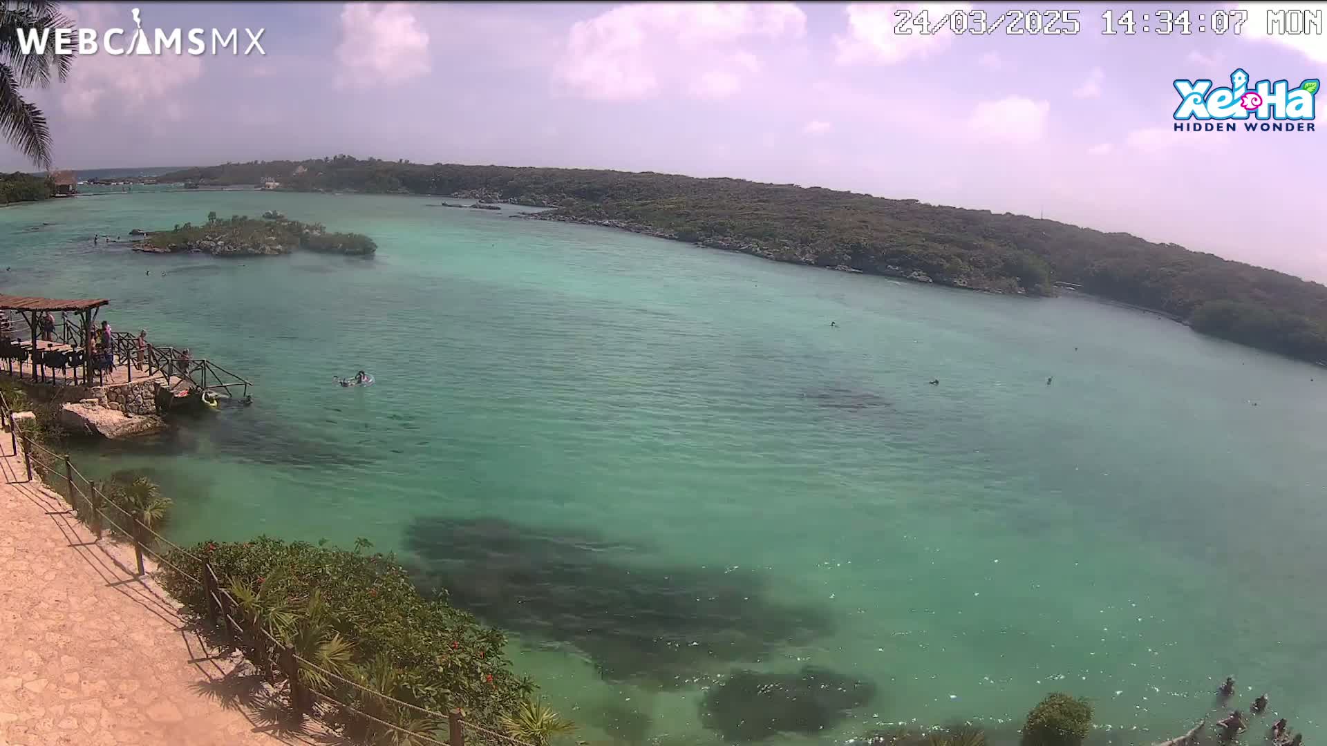 A partly sunny day shows a calm, clear, turquoise body of water with several people swimming and wading near a rocky shoreline with vegetation.