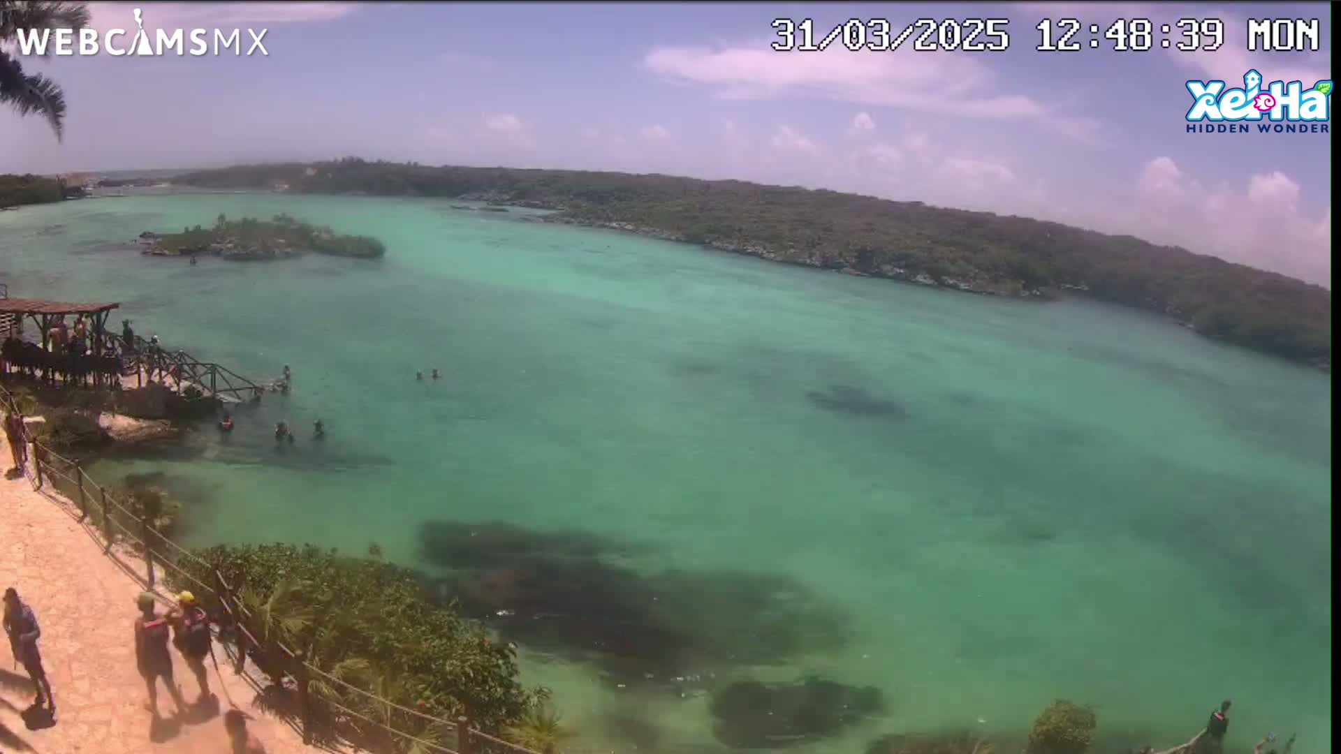A mostly sunny day reveals a clear, bright teal body of water with people swimming and relaxing near a rocky shore and lush green foliage.