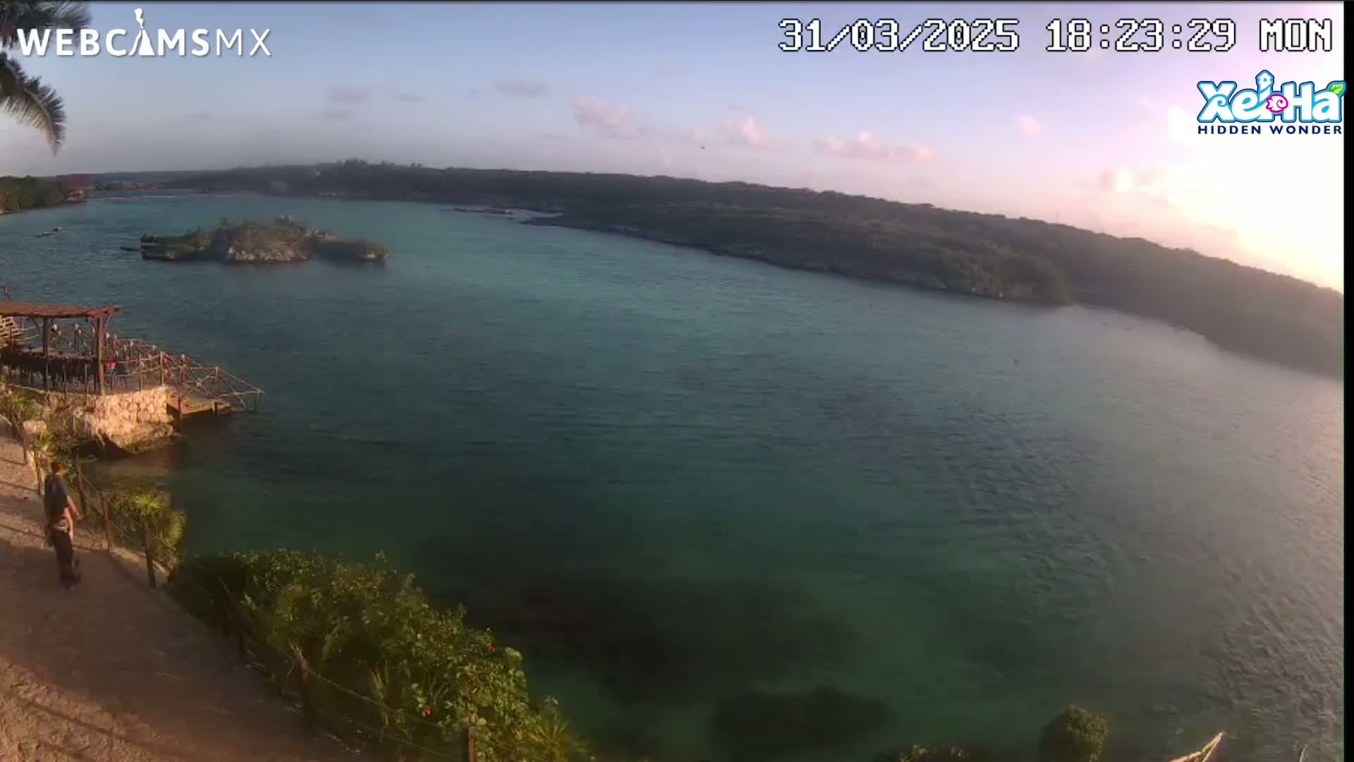 A calm, teal-colored bay is seen from an elevated vantage point at sunset, with a small island visible in the middle distance and a person standing near a wooden structure on the shore.