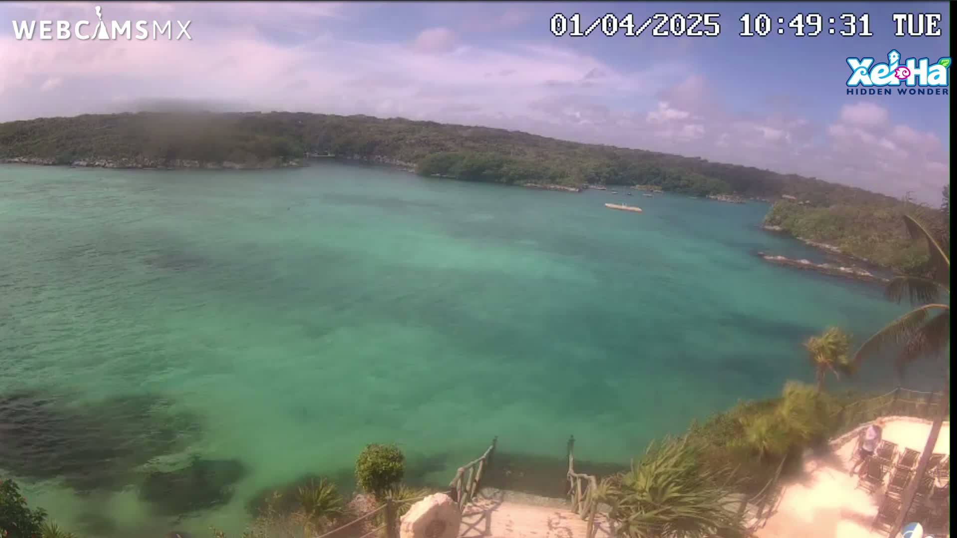 A high-angle view shows a calm, clear, turquoise body of water surrounded by lush green vegetation under a partly cloudy sky.