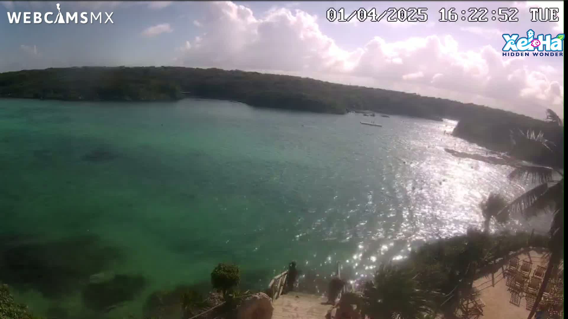 A partly sunny day shows a clear, bright green body of water partially enclosed by land, with a staircase leading down to it from a palm tree-lined shore.