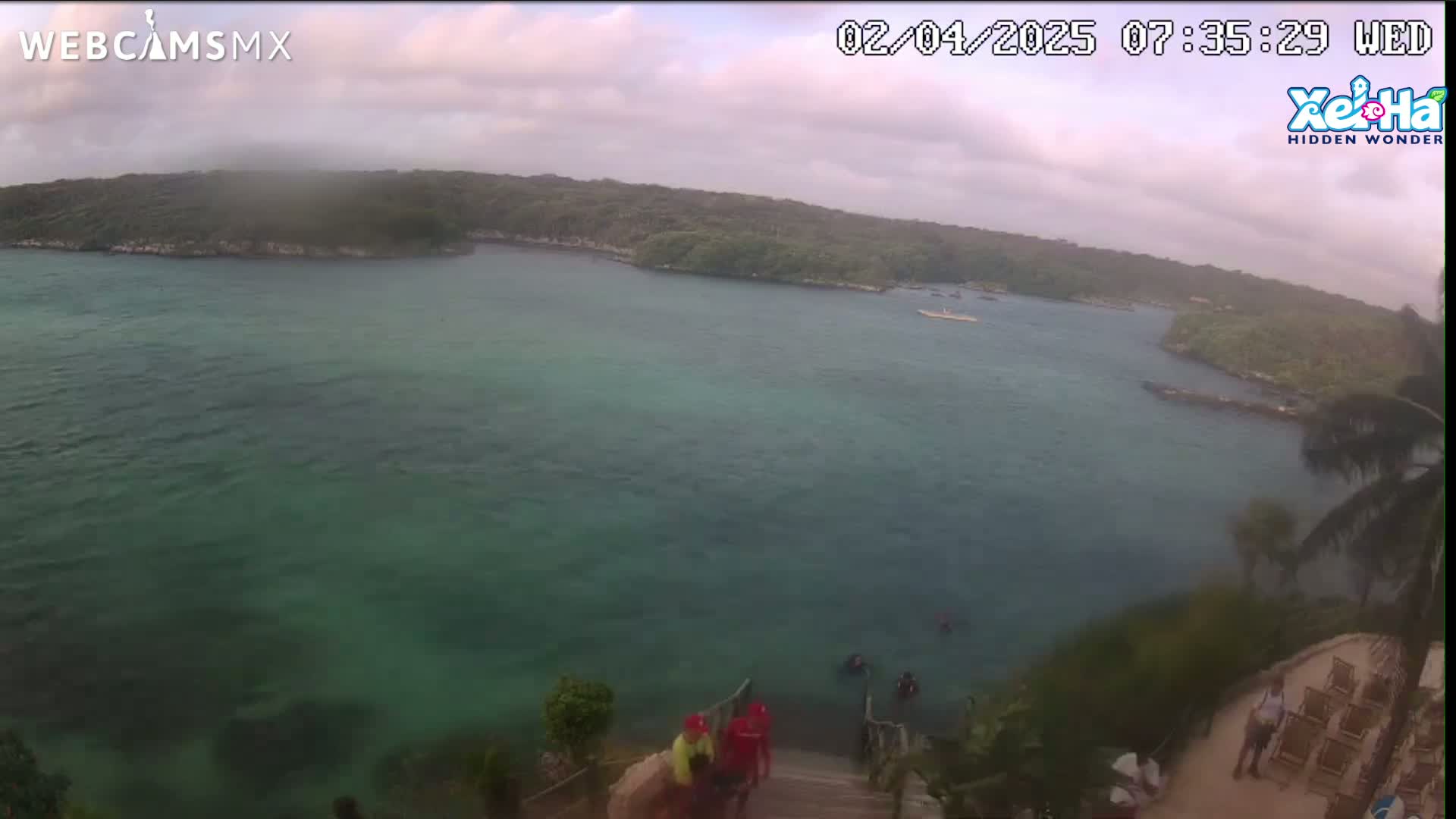 A tranquil, overcast day reveals a calm body of teal water nestled between lush green hillsides, with several people near a wooden staircase leading down to the water's edge.