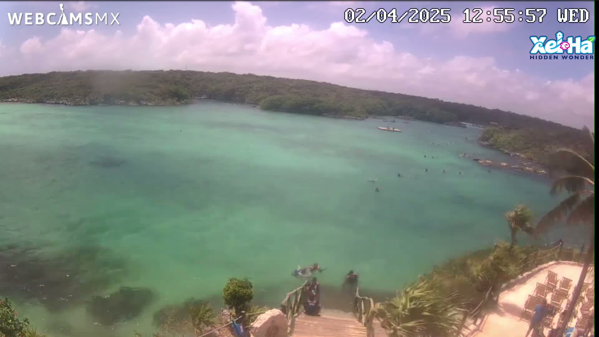 A partly cloudy day reveals a tranquil cove of bright teal water, with several people swimming and a wooden staircase leading down to the water's edge from a higher vantage point.