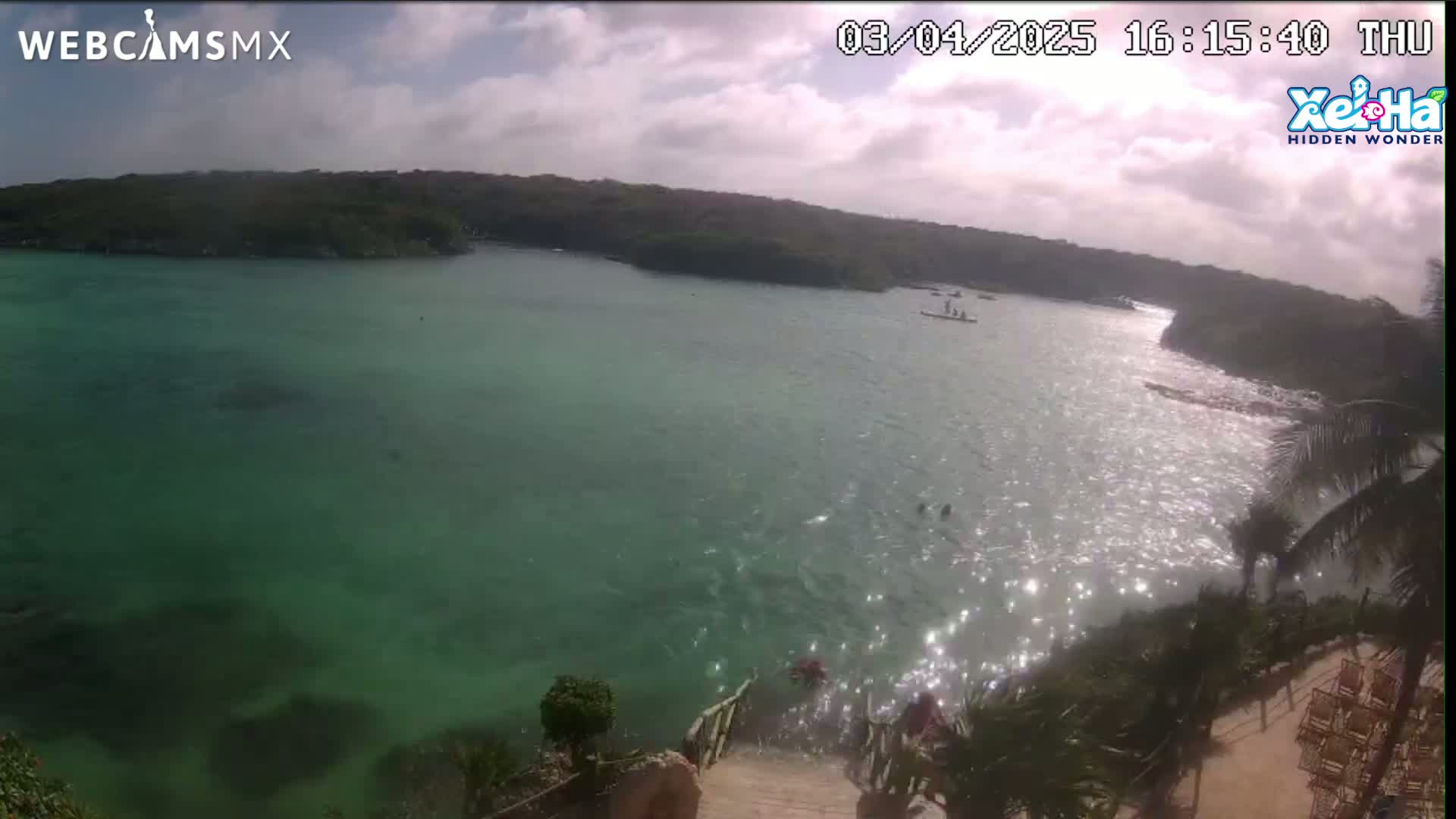 A partly sunny day shows a tranquil bay of clear, green water with people swimming and some small boats, surrounded by lush green vegetation and a stone staircase leading down to the water's edge.