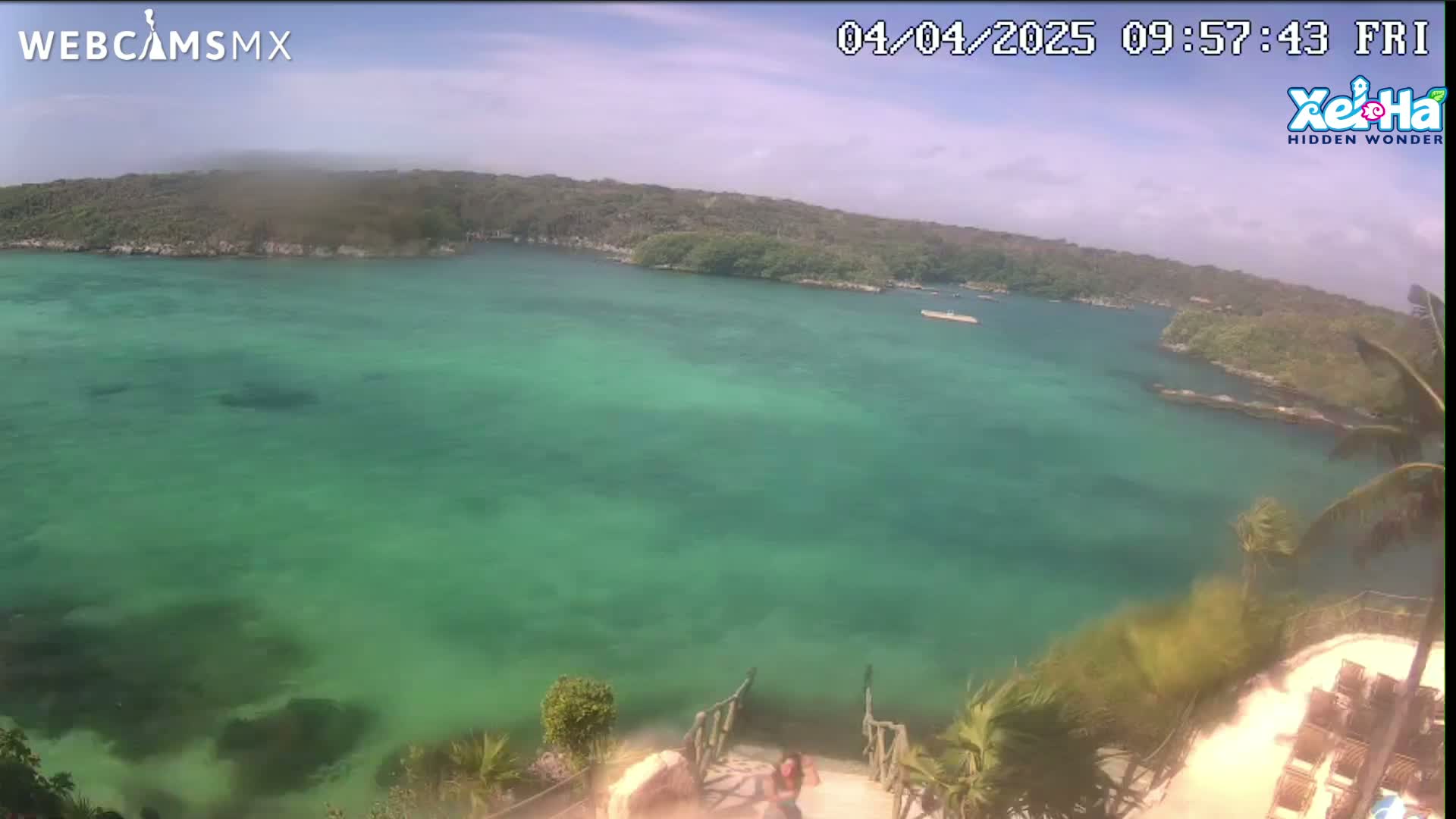 A high-angle view shows a calm, clear, teal-green body of water surrounded by lush green vegetation under a mostly sunny sky.
