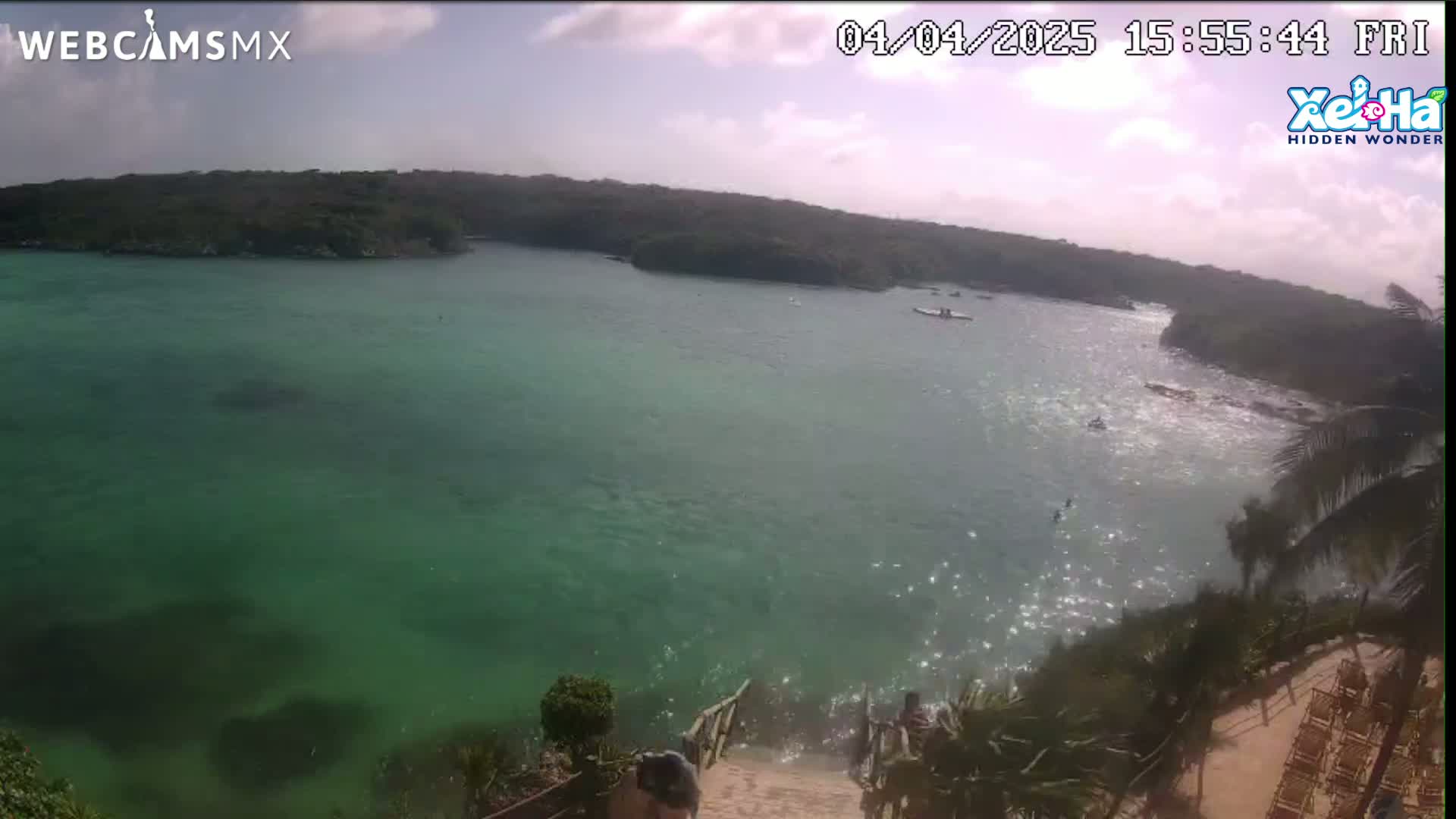 A tranquil, bright, sunny day reveals a view of a calm, clear turquoise body of water surrounded by lush green vegetation, with several small boats visible in the distance.
