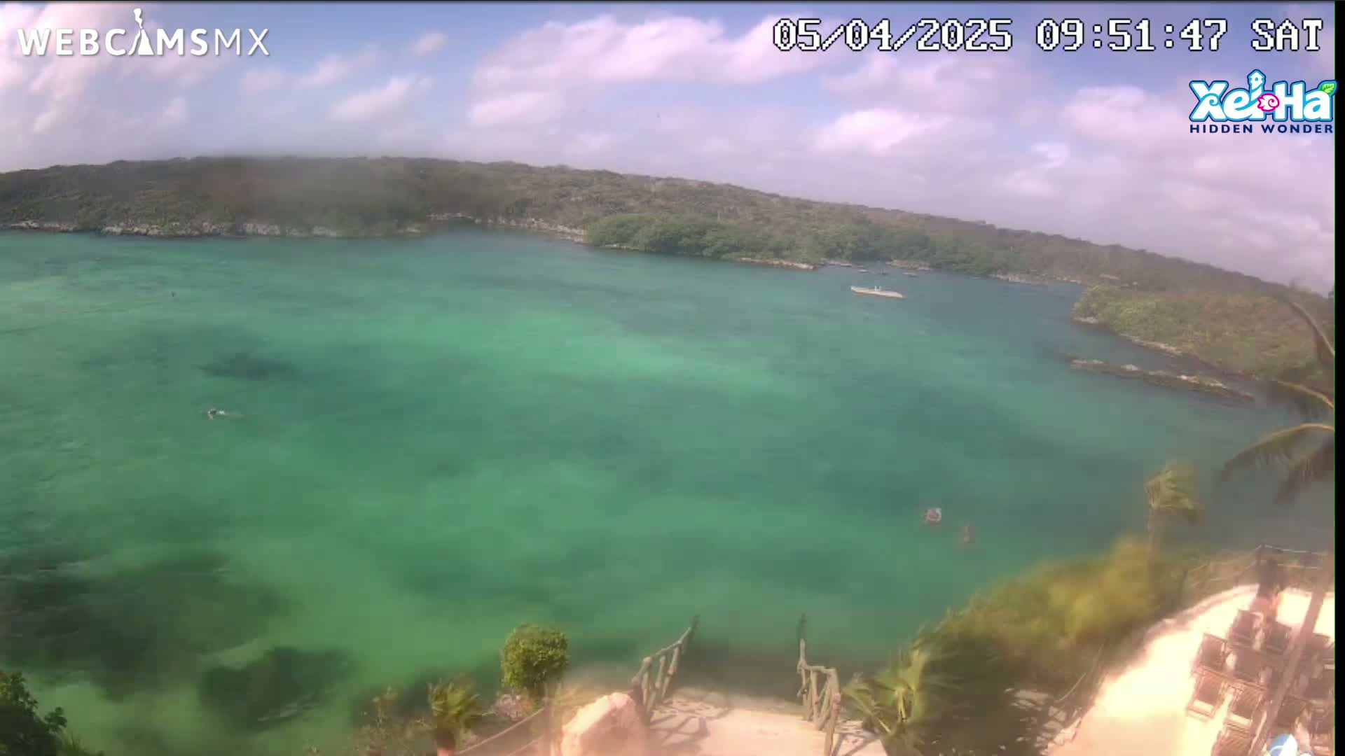 An aerial view shows a calm, bright green body of water partially surrounded by lush green vegetation under a partly cloudy sky.