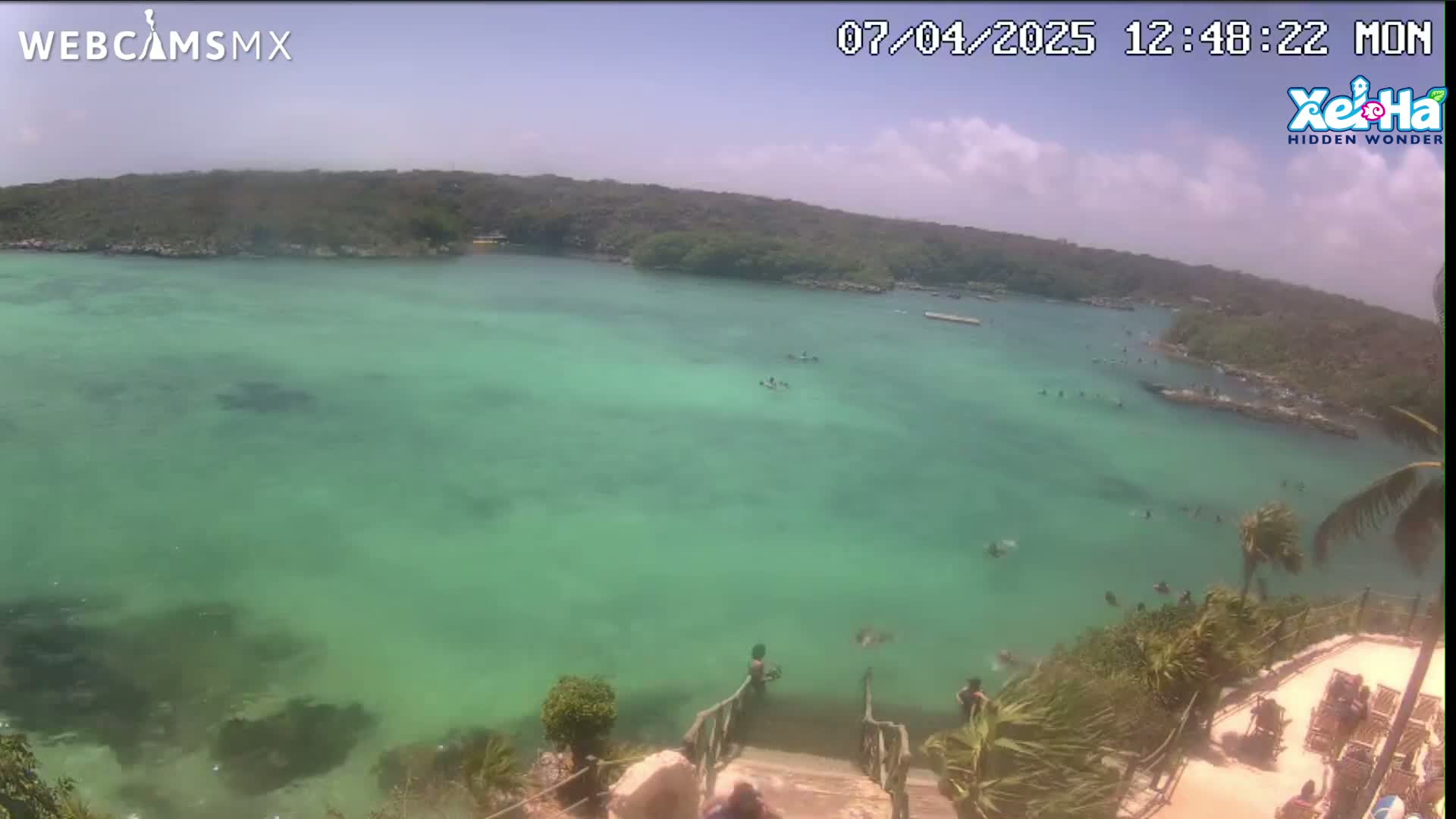 A high-angle view shows a calm, clear, teal-colored body of water with people swimming and lounging near a rocky shoreline on a partly sunny day.