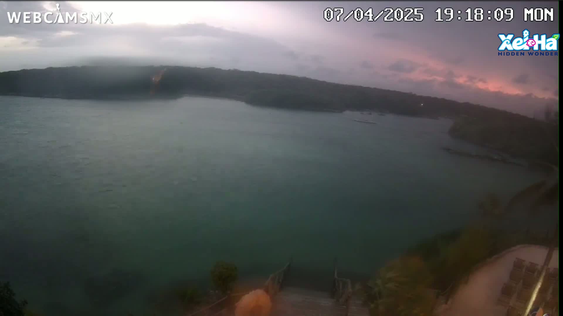 A calm body of water is seen from an elevated viewpoint under a cloudy, overcast sky with hints of pink near the horizon.