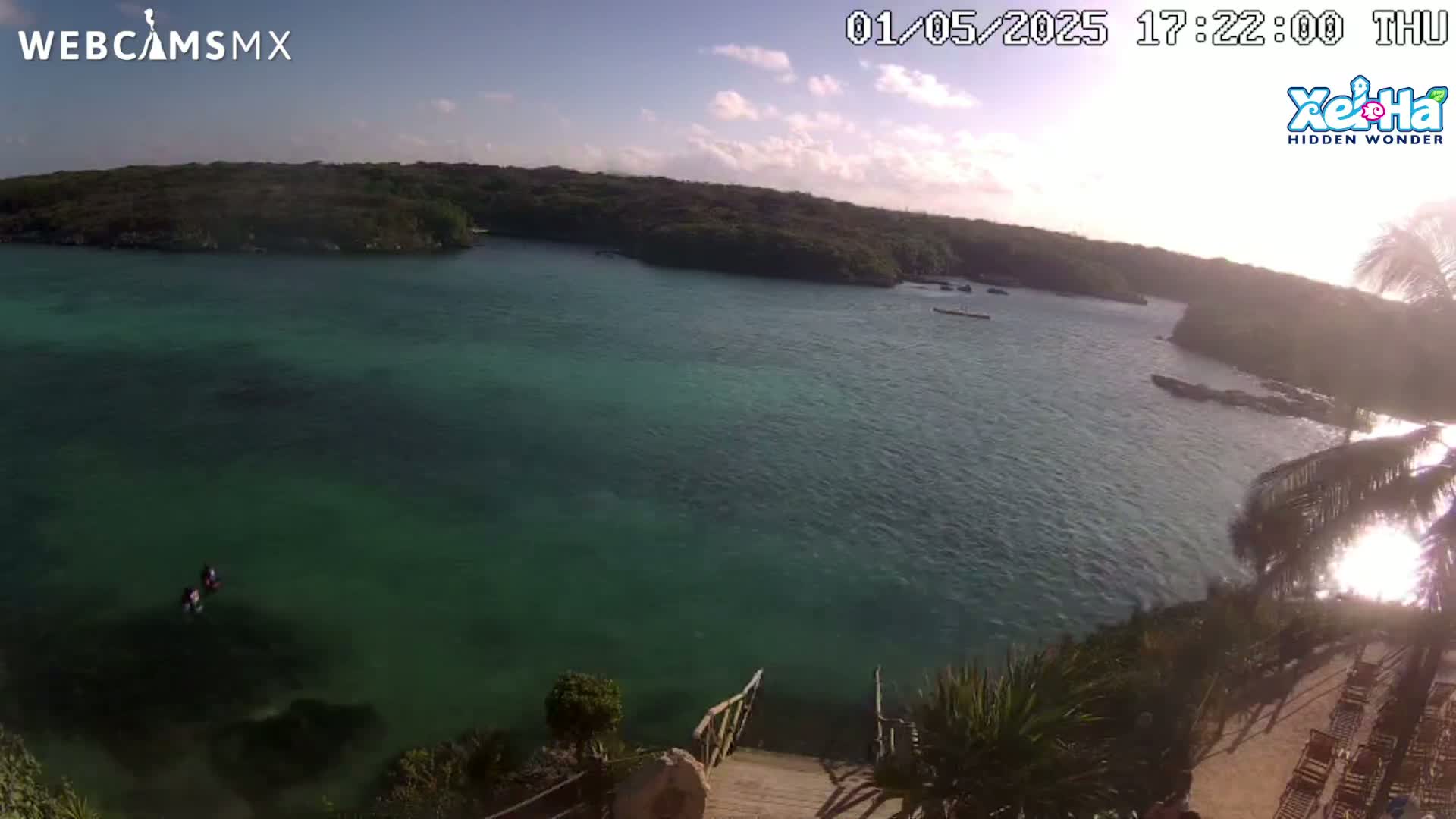 A tranquil, clear teal-green bay is nestled between verdant hills under a sunny, mostly clear sky, with a few boats visible in the distance and two people swimming in the shallows.