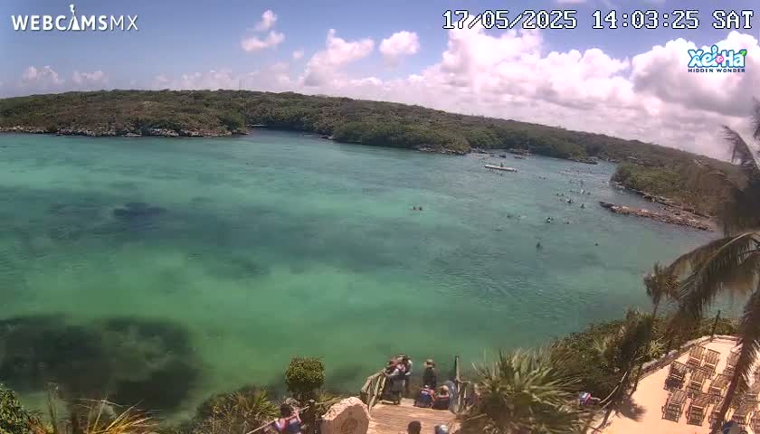 A high-angle view shows a calm, clear, teal-colored body of water surrounded by lush green vegetation under a partly cloudy sky.