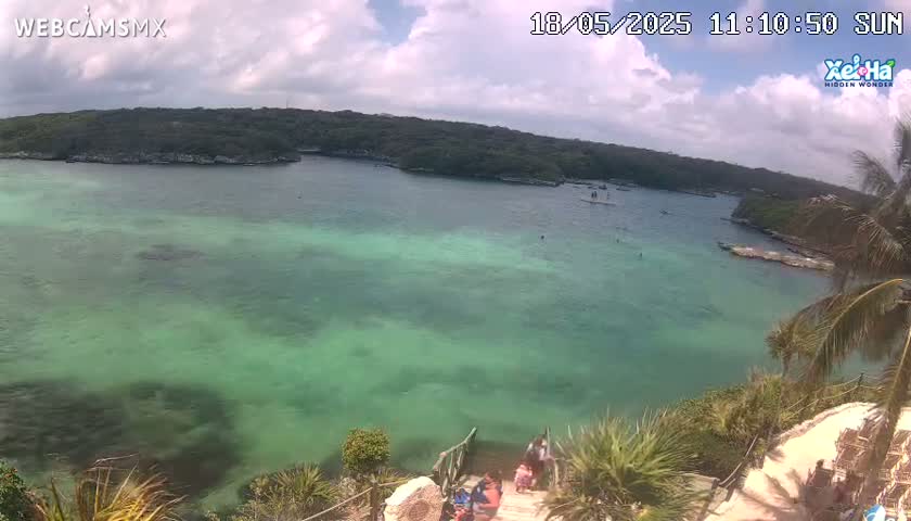 A partly sunny day shows a tranquil, shallow, teal-green body of water surrounded by lush green vegetation, with several people visible near a set of stairs leading down to the water.