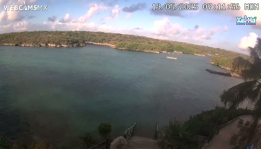 A partly cloudy sky overlooks a calm body of teal water surrounded by lush green vegetation, with a wooden staircase leading down to the water's edge.