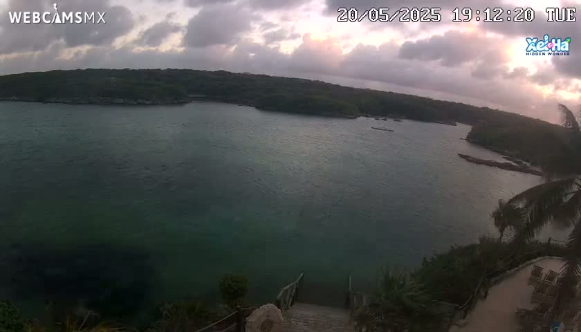 A tranquil cove with calm, dark-green water is nestled within a lush, green landscape under a cloudy, dusky sky.