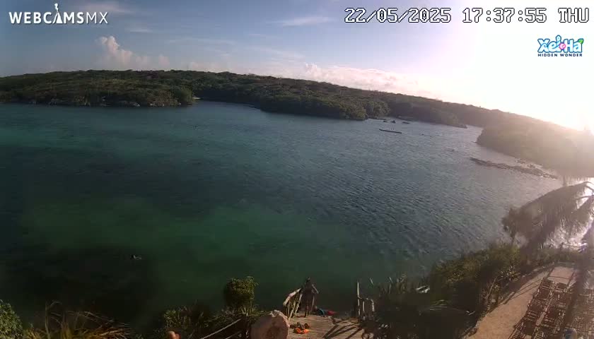A high-angle view shows a calm, teal-colored body of water nestled between lush green hills under a mostly clear, sunny sky.