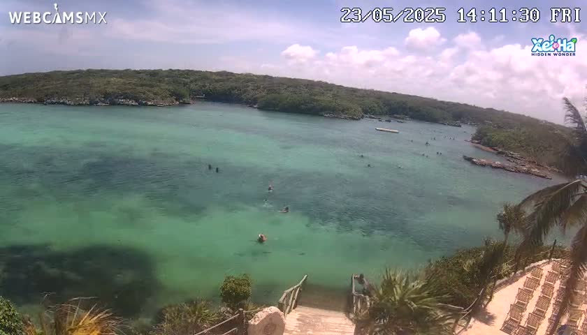 A partly cloudy day shows a tranquil, shallow, green-blue body of water with several people swimming, surrounded by lush green vegetation and a stairway leading down to the water's edge.