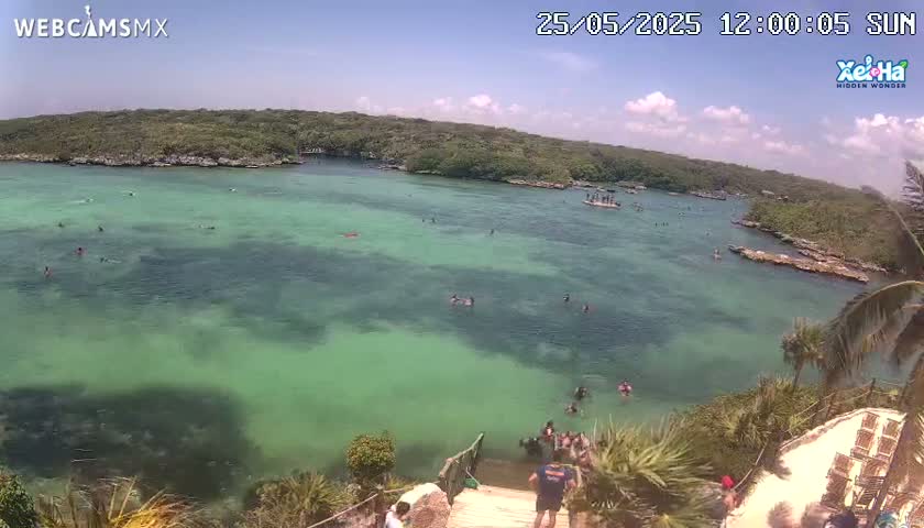A high-angle view shows a calm, clear, turquoise body of water with people swimming and wading, surrounded by lush green vegetation under a sunny sky with a few clouds.