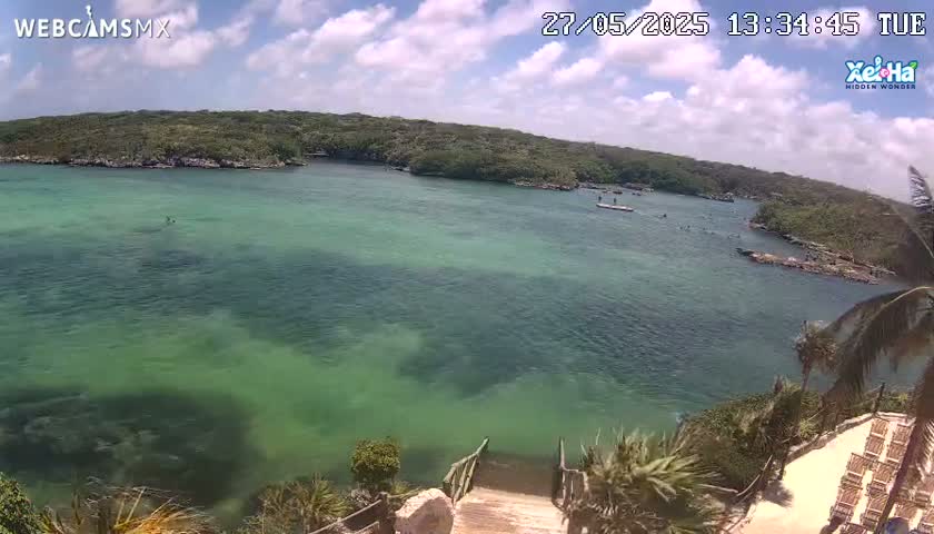 A partly cloudy day shows a tranquil, shallow, teal-colored body of water surrounded by lush green vegetation and a wooden staircase descending to the water's edge.