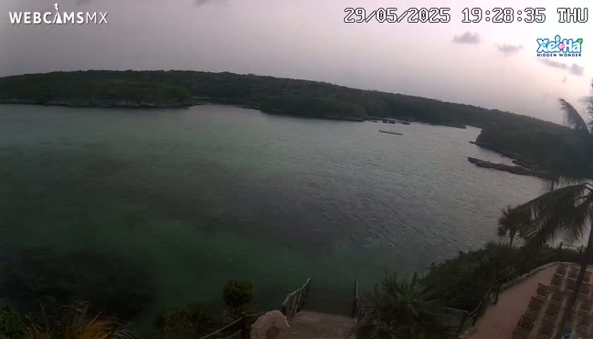 An overcast sky hangs over a calm, teal-green bay nestled between lush, green hillsides, with wooden stairs descending to the water's edge.