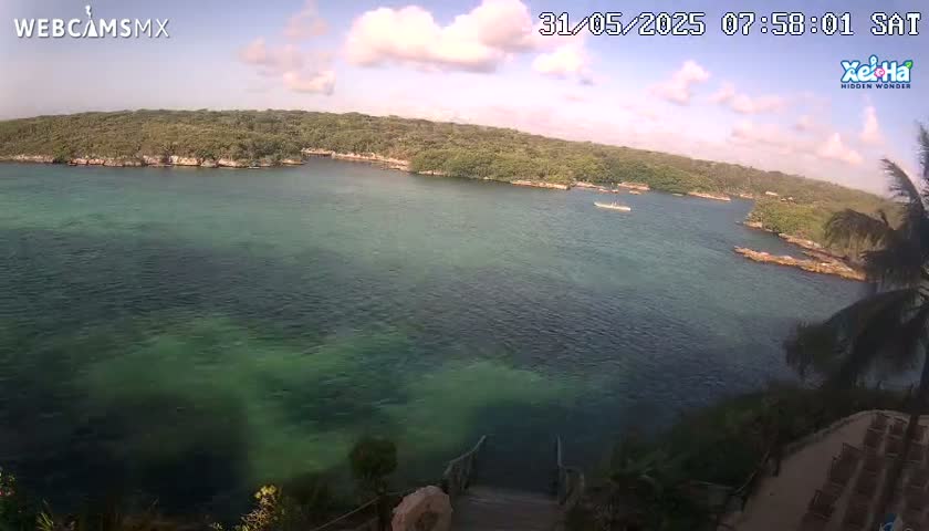 A partly cloudy day reveals a tranquil bay of clear, teal water partially surrounded by lush green vegetation, with a small boat visible in the distance.