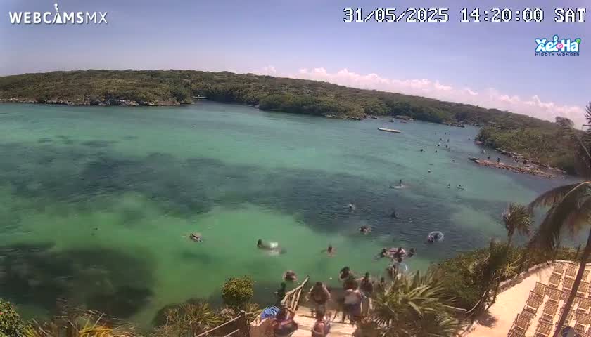 An aerial view shows a calm, clear, teal-green body of water with people swimming and lounging near a rocky shore on a sunny day.