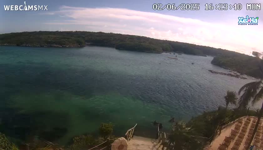 A tranquil bay with calm water of varying shades of green and blue is viewed from a high vantage point, under a mostly sunny sky with a few clouds.