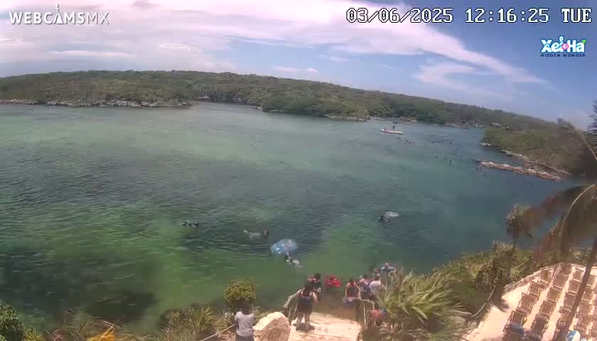 A calm, partly cloudy day overlooks a tranquil bay with clear, shallow green water where people are swimming and wading near a rocky shore with lush vegetation and wooden chairs.