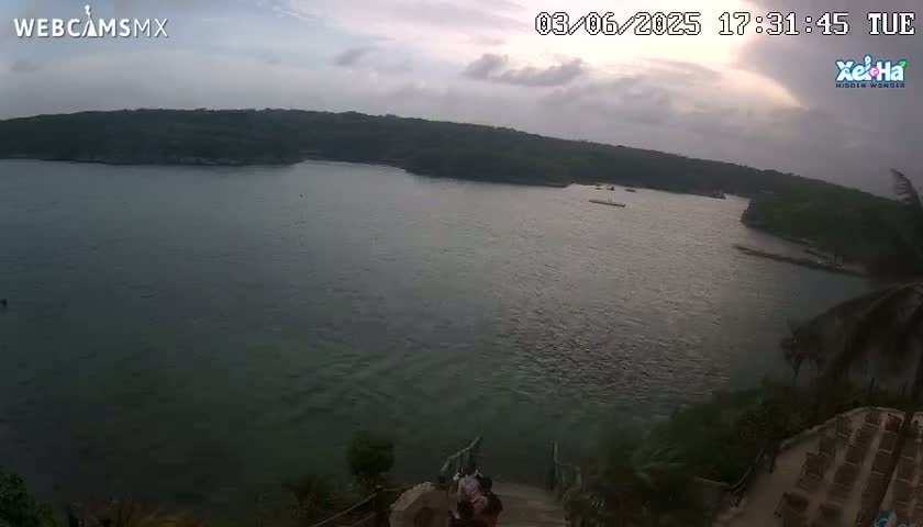 An elevated view overlooking a calm body of water under a mostly cloudy sky, with a few people visible near a staircase leading down to the water's edge.