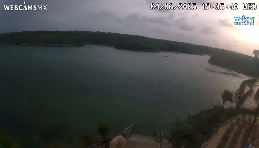 An overcast sky hangs over a calm bay, partially surrounded by lush green hillsides, with a wooden staircase leading down to the water's edge from a partially visible structure.