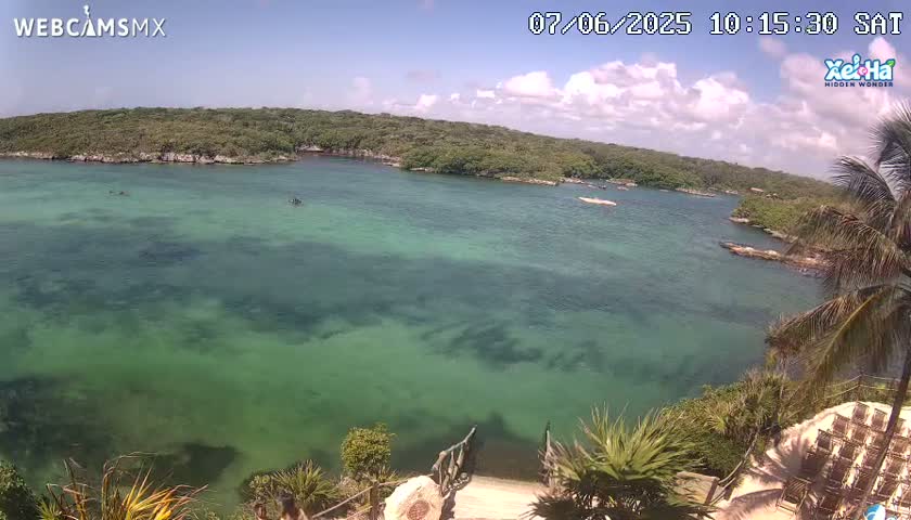 A tranquil, clear, green-blue body of water is surrounded by lush green vegetation under a partly sunny sky.
