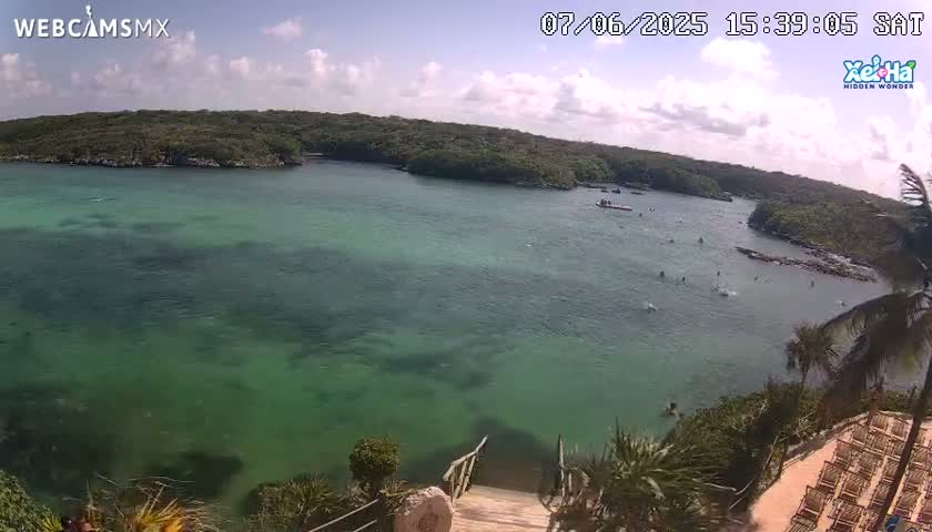 A partly cloudy day shows a calm, green-blue body of water surrounded by lush green vegetation, with several people swimming and wading in the water.
